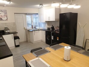 Une cuisine compacte avec un réfrigérateur noir, un lave-linge et un coin repas comprenant une table et des chaises en bois.