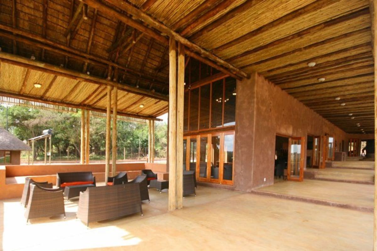 An open-air structure with a wooden ceiling and large windows, furnished with benches and tables.
