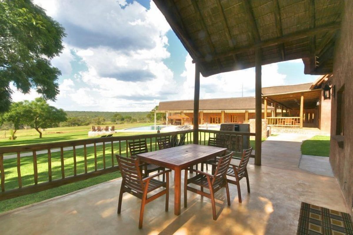 An outdoor patio with a wooden table and chairs, a covered area, and a view of a building and landscape.