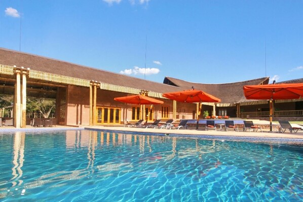 A swimming pool with lounge chairs and umbrellas, surrounded by a building with a thatched roof.