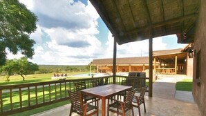 Eine Terrasse im Freien mit einem Holztisch und Stühlen, einem überdachten Bereich und Blick auf ein Gebäude und die Landschaft.
