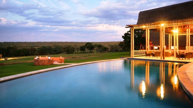A swimming pool with a wooden deck and lounge area, surrounded by a grassy landscape and a distant hill. - Olievenfontein Private Game Reserve (Vaalwater)