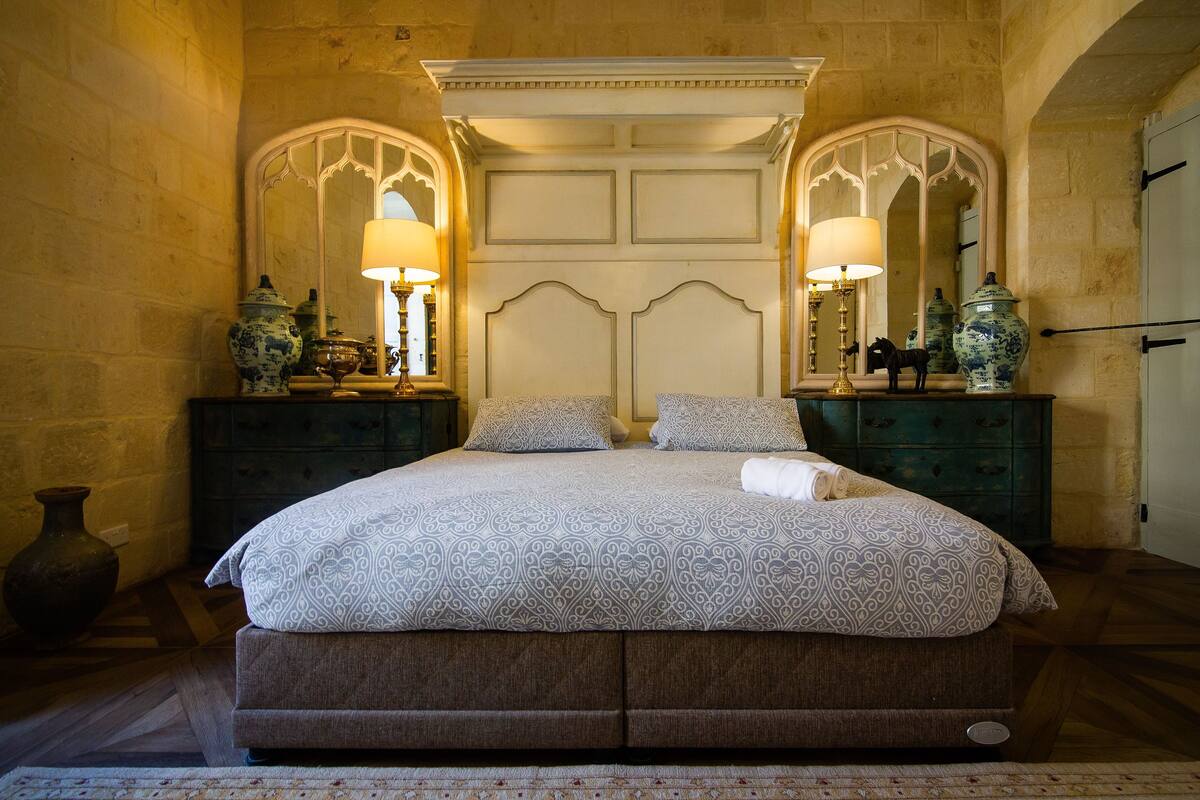 Una cama grande con una colcha estampada, dos mesitas de noche con lámparas y un espejo sobre la cama.