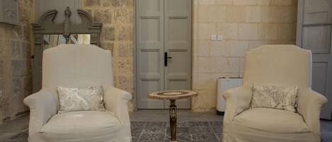 Dos sillones con cojines estampados, una mesita redonda y una alfombra con diseño en una habitación con paredes de piedra y una puerta al fondo.