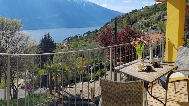 Un balcón con vistas a un lago y montañas, mobiliario de mimbre y una mesa con tazas y un florero.