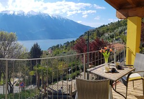 Varanda com vista para um lago e montanhas, mobiliário de vime e uma mesa com chávenas e um vaso de flores.
