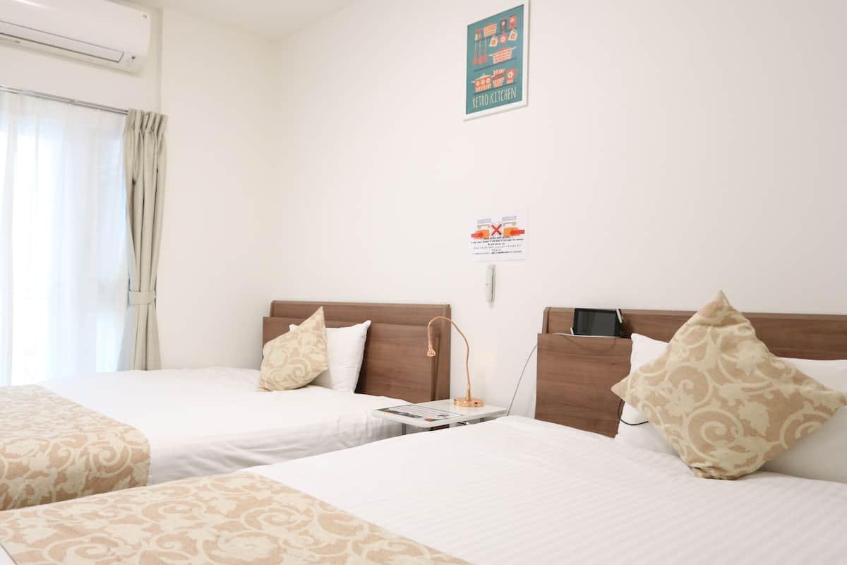 A hotel room with two beds, a bedside table with a lamp, a wall-mounted air conditioner, and a poster on the wall.