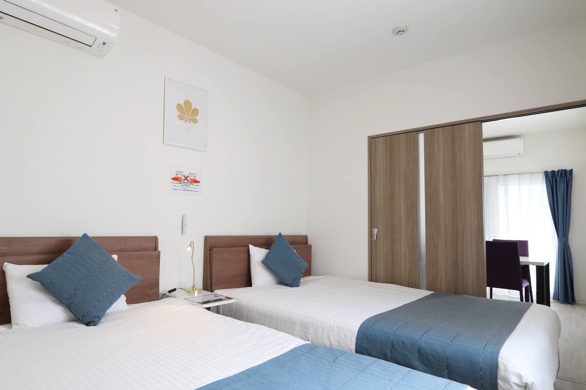 A hotel room with two beds, a bedside table with a lamp, a wall-mounted air conditioner, and a door leading to a balcony with a view.
