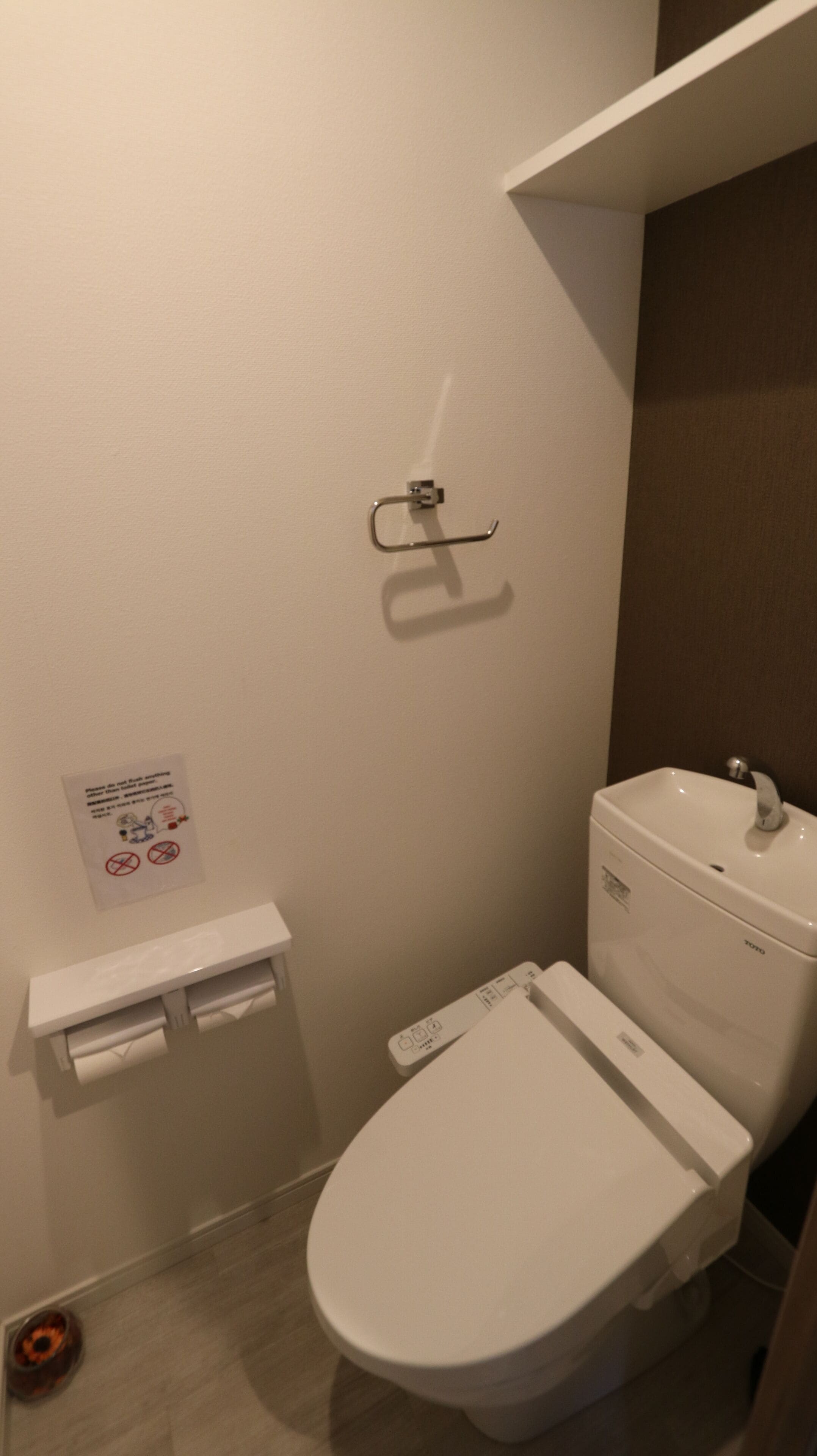 Uma casa de banho pequena e limpa, com sanita, lavatório e secador de mãos. Há um aviso na parede com o símbolo de proibido fumar e um alerta sobre o chão molhado.
