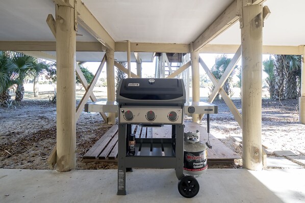 A portable barbecue grill with a propane tank underneath, set up on a wooden deck with a palm tree in the background.