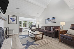 A spacious living room with a high ceiling, a large window, and a view of the outdoors. There is a brown leather sofa, a wooden coffee table, and a blue star-patterned rug. - 2 BR + Loft/2BA Tarpon Pond Cottage! Great Views! Amenity access included (Seabrook Island)