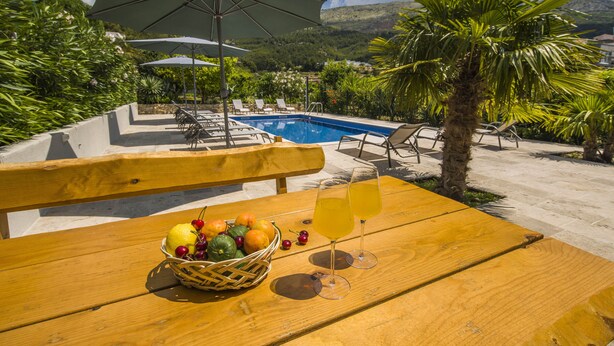 Taula de fusta amb una cistella de fruita i dues copes de beguda groga, una piscina, gandules i ombrelles en un entorn de resort.