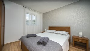 Un llit ben fet amb un edredó gris, una taula de nit de fusta amb una làmpada, i una finestra amb cortines translúcides.