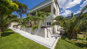 Una casa moderna de dos pisos amb un balcó cobert, envoltada de vegetació abundant i un cel blau clar.
