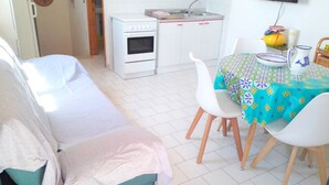 Een kleine, schone kamer met een bed, een tafel met stoelen, een keukenhoek met fornuis en gootsteen, en een klok aan de muur.
