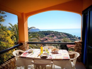 Tavolo da balcone apparecchiato con piatti, posate e una bottiglia, con vista su una cittadina costiera e una limpida visuale del mare e delle montagne.