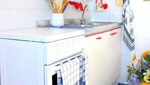 Een kleine keuken met witte kastjes, een inbouwoven en diverse keukengerei dat boven de aanrecht hangt.