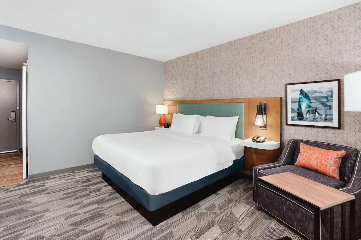 A hotel room with a large bed, a bedside table with a lamp, a chair with a cushion, and a framed picture on the wall.