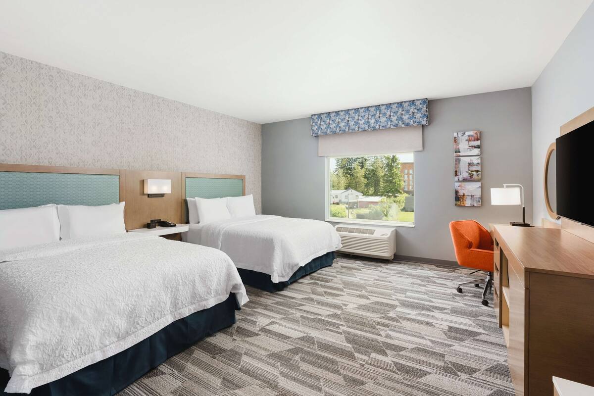 A hotel room with two beds, a desk, an orange chair, a large window, and a flat-screen TV.