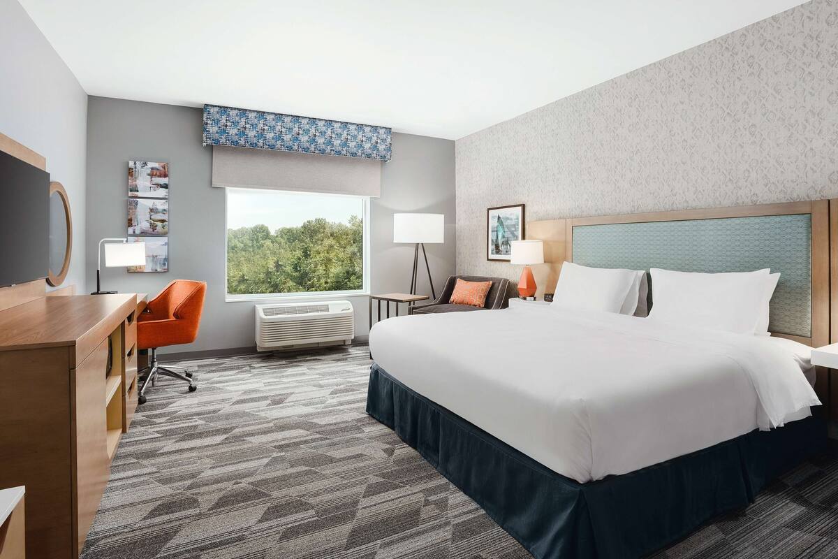 A hotel room with a large bed, a desk, an orange chair, a window with a view of trees, and a patterned carpet.