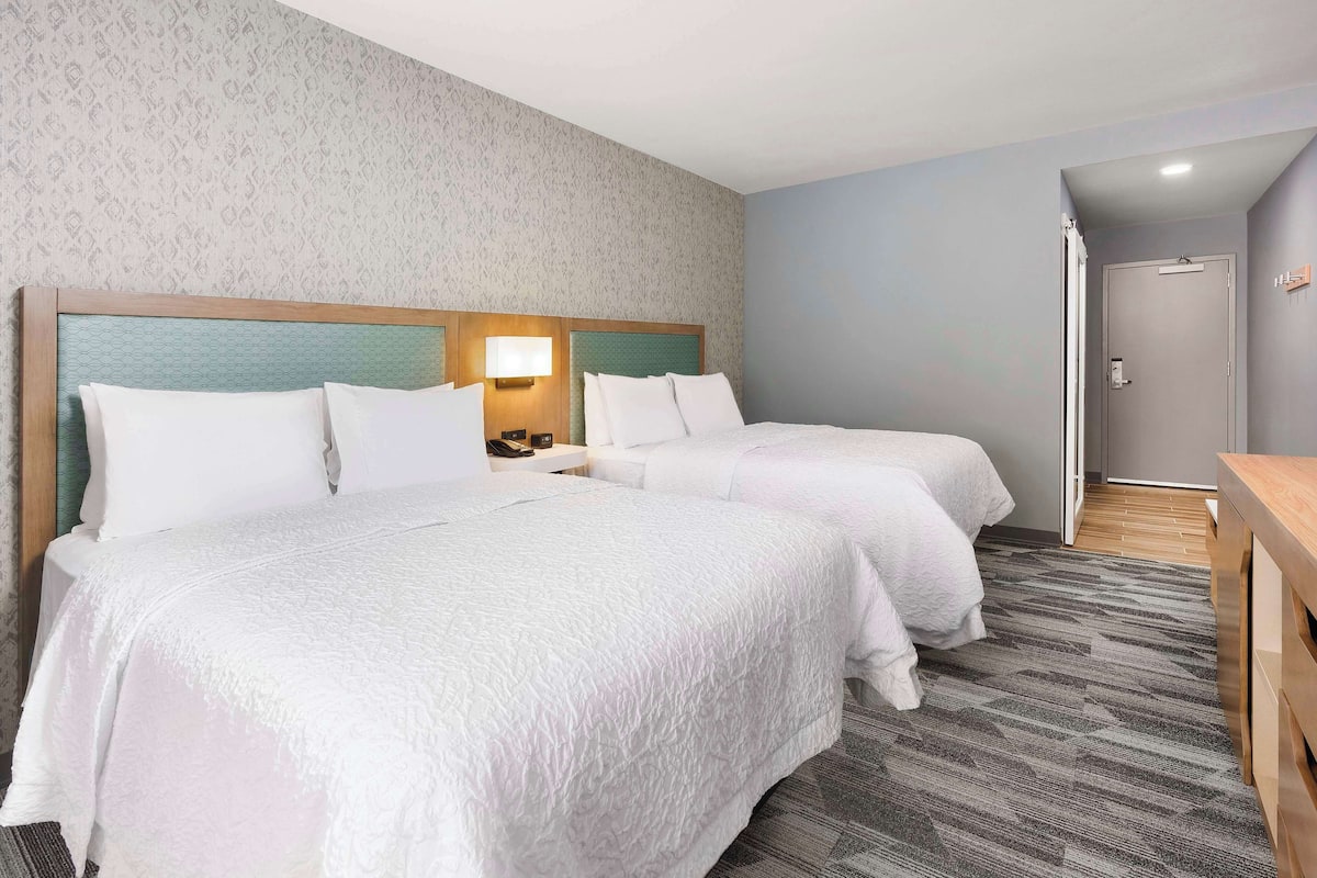 A hotel room with two beds, a nightstand, a lamp, and a door.