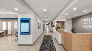 A hotel reception area with a digital departure board, a reception desk, and a "Welcome to the Event Green" sign.