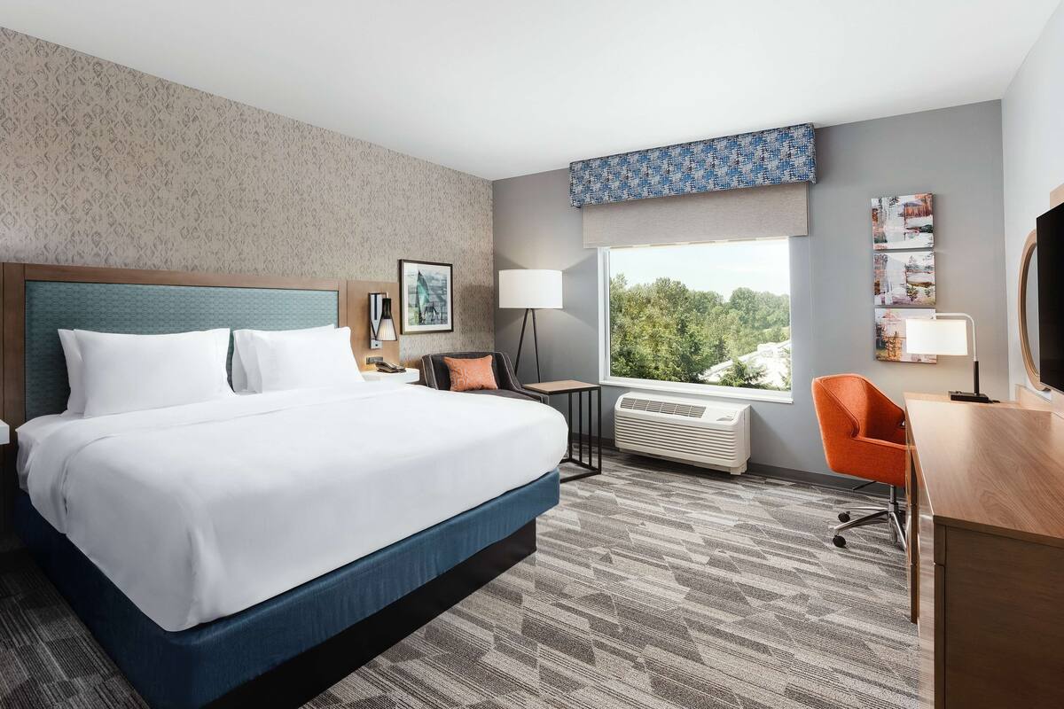 A hotel room with a large bed, a desk, an orange chair, a window with a view of trees, and a wall with patterned wallpaper.