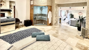 Une chambre d’hôtel moderne avec un grand lit, un coin salon agrémenté d’un tapis à motifs, un espace dédié aux massages avec un mur en pierre, et un comptoir de réception en arrière-plan.