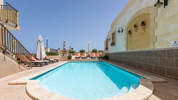 Une piscine avec des chaises longues et des parasols, un escalier et un bâtiment sur lequel est affiché un panneau indiquant « INTERDIT DE PLONGER ».