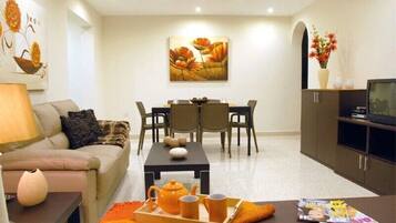 Un salon moderne avec un canapé, une table basse et un coin repas. On y trouve un téléviseur posé sur un meuble et un tableau accroché au mur.