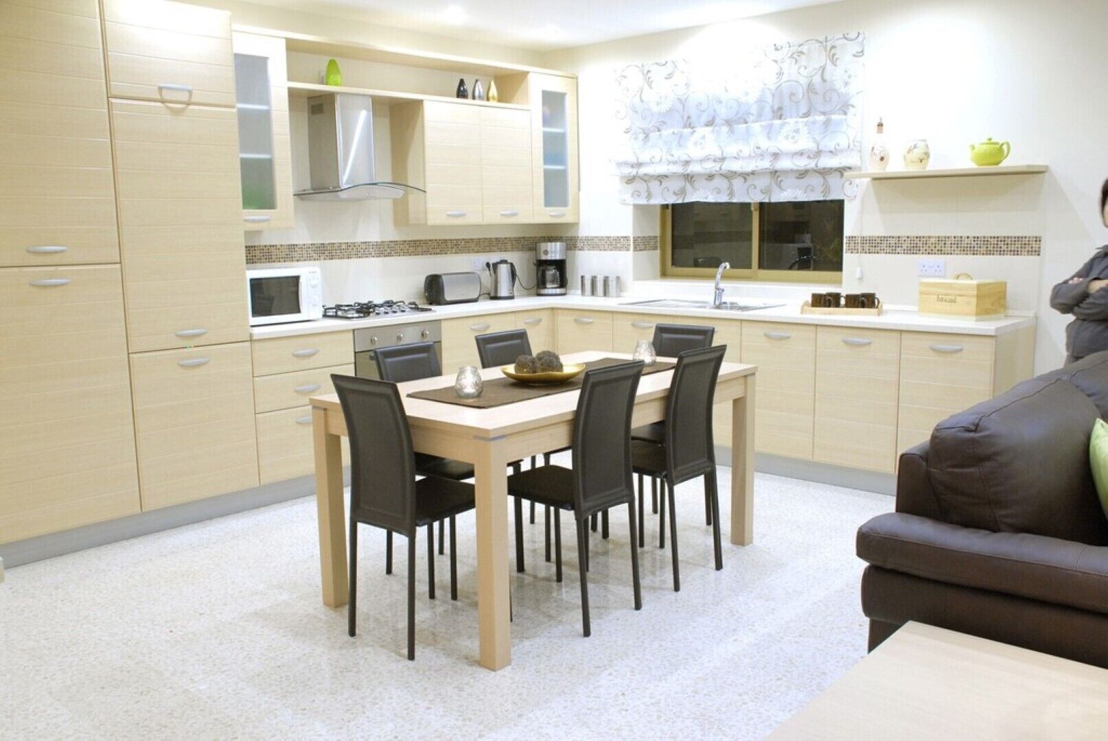 A modern kitchen with light-colored cabinets, a dining table with chairs, and a brown sofa in the background.