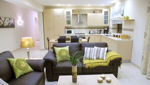 Een woonkamer met een bank, salontafel en een eethoek met stoelen, met een keuken op de achtergrond.