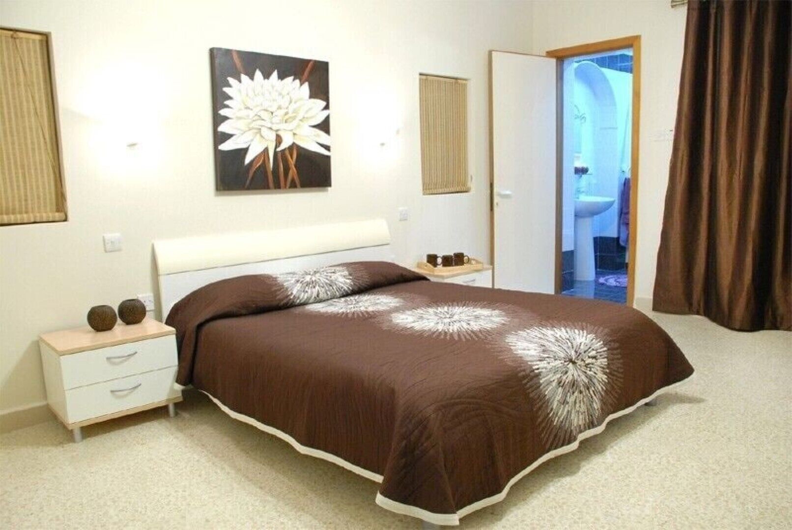 A bedroom with a large bed, a bedside table with two decorative items, a wall-mounted painting, and a door leading to a bathroom.