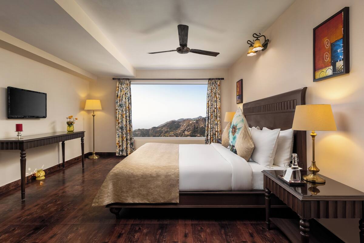 A hotel room with a bed, a ceiling fan, a TV, a window with curtains, and a painting on the wall.