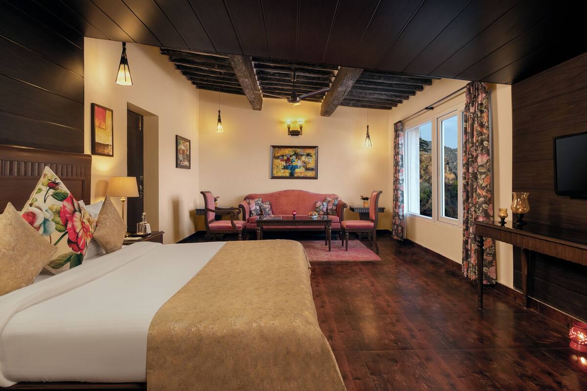 A hotel room with a large bed, a sofa, a coffee table, and a TV. There are windows with curtains, a ceiling with exposed beams, and artwork on the walls.
