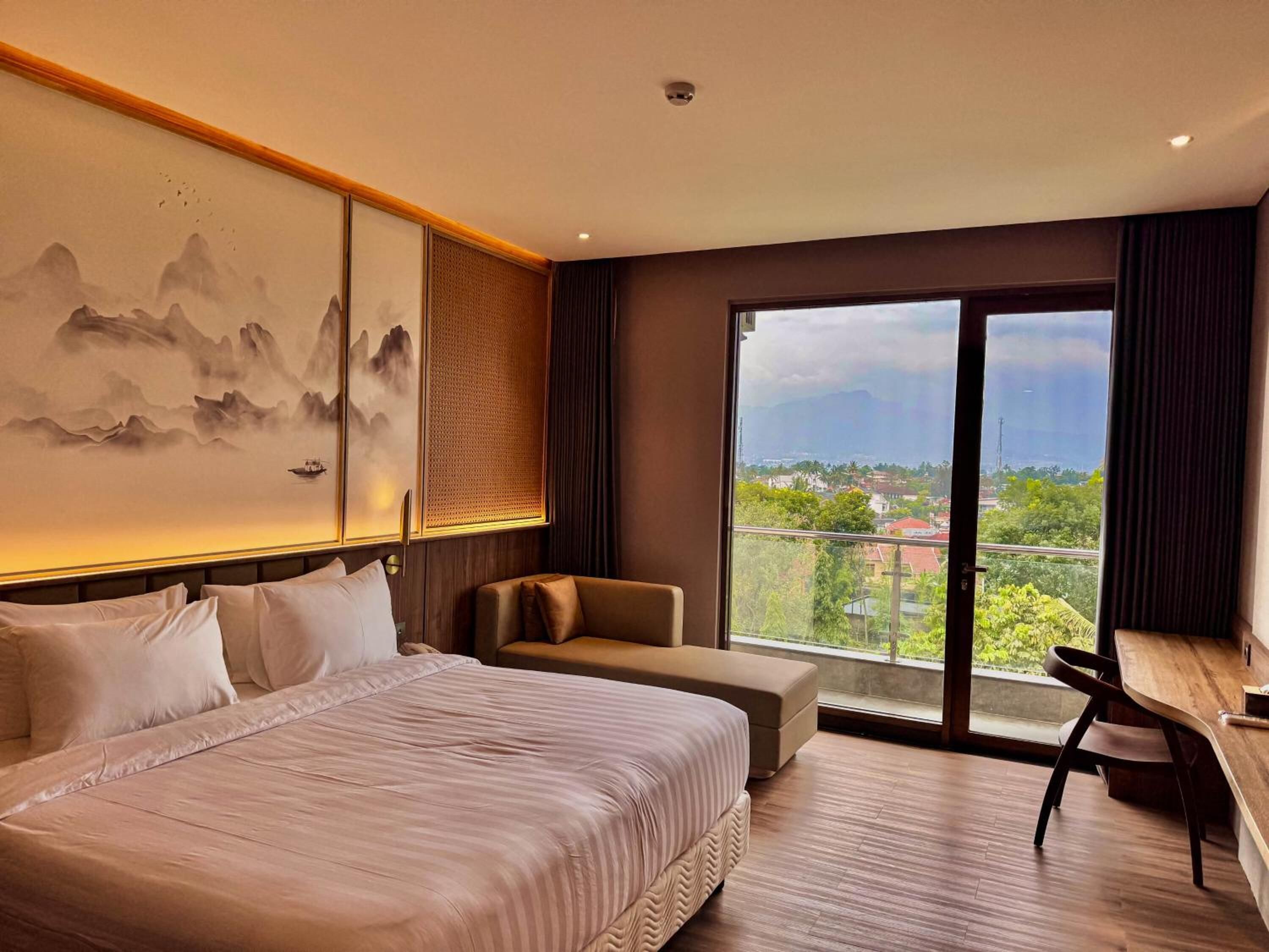 A hotel room with a large bed, a sofa, a desk, and a view of a cityscape through a large window.