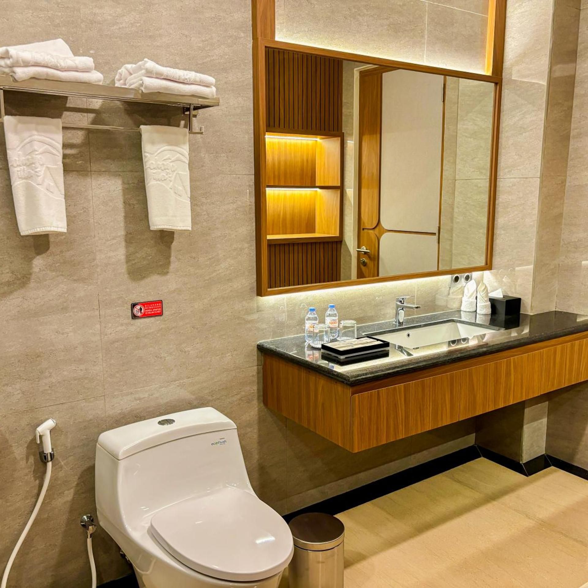 Ein modernes Badezimmer mit Toilette, Waschbecken und Spiegel. Handtücher befinden sich an einem Wäscheständer, und ein Schild ist an der Wand angebracht.