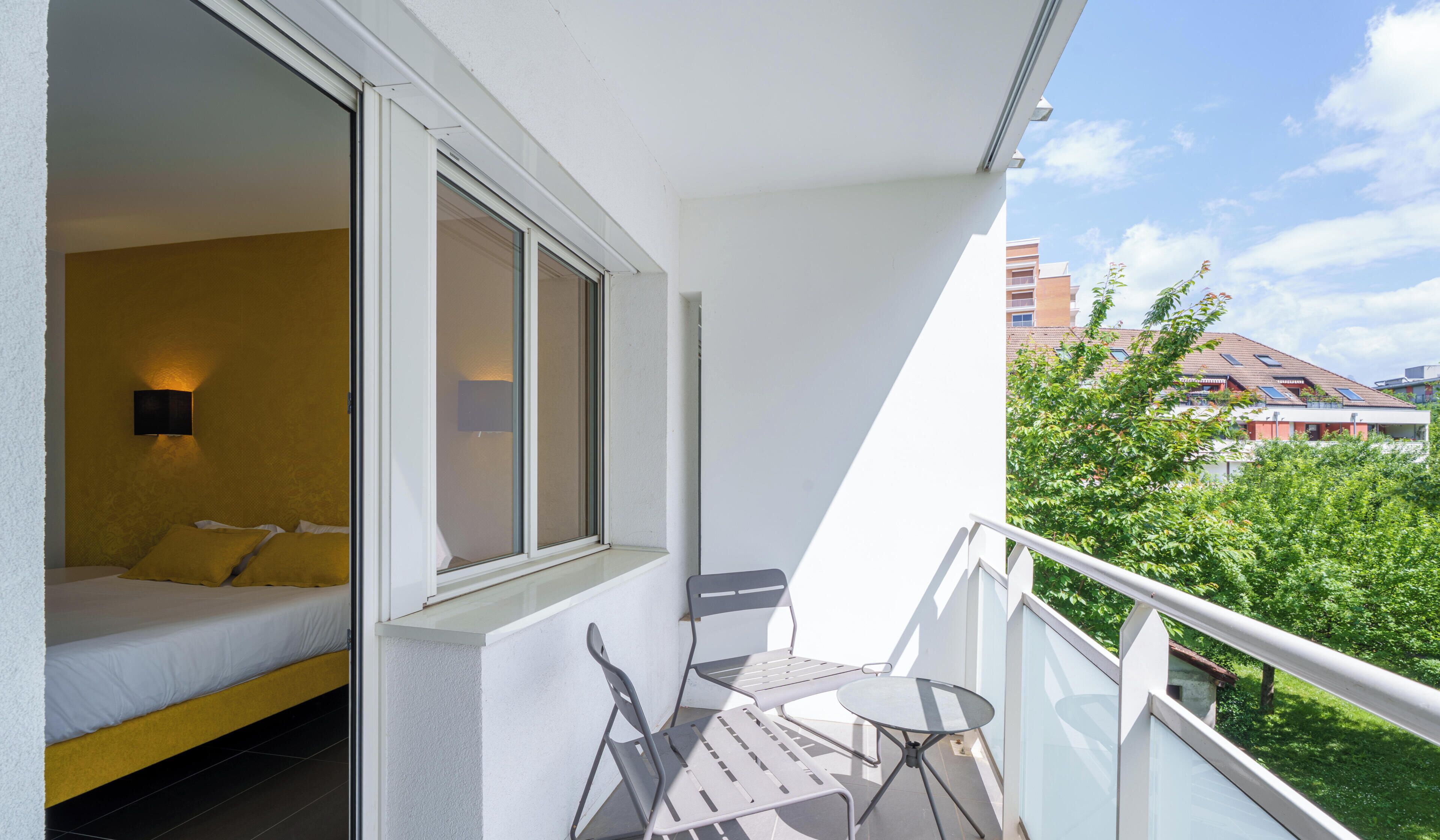 A balcony with a bed, a table, and chairs, overlooking a green area and buildings.