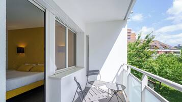 A balcony with a bed, a table, and chairs, overlooking a green area and buildings.