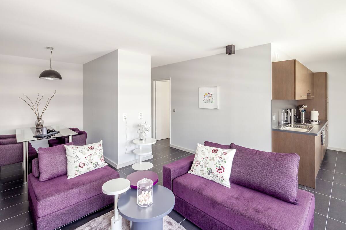 A modern living room with two purple sofas, a small round table, and a kitchen area in the background.