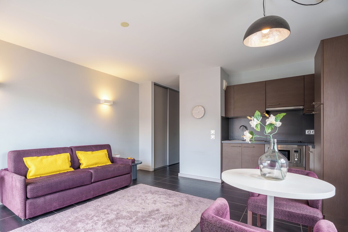 A modern living room with a purple sofa, a white table, and a kitchen area with dark cabinets.