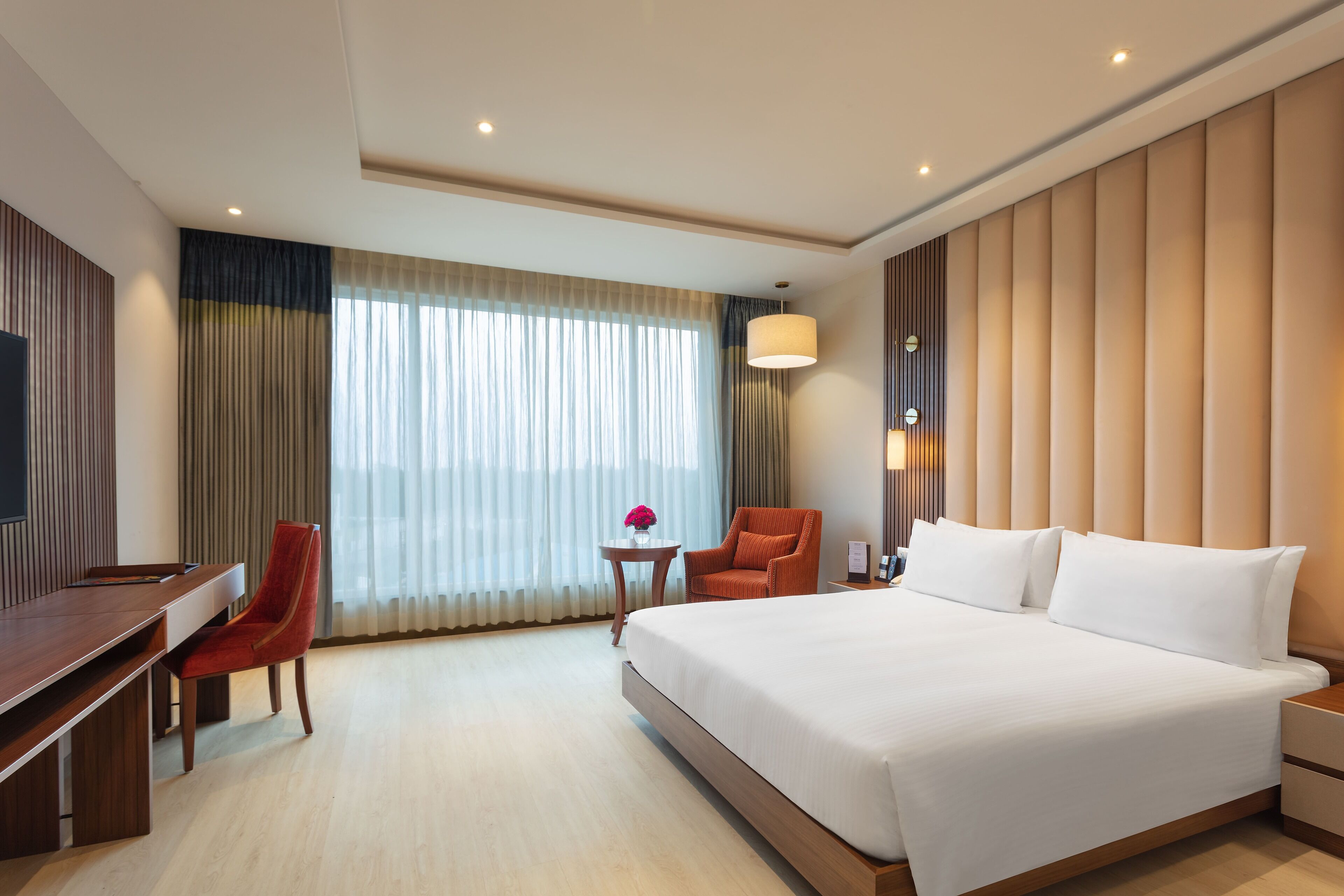 A hotel room with a large bed, a desk, a chair, a small table with a vase, and a large window with curtains.