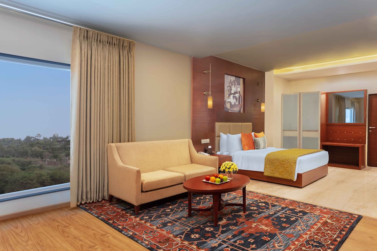 A hotel room with a sofa, a bed with a yellow blanket, a bedside table with fruit, a patterned rug, and a large window with curtains.