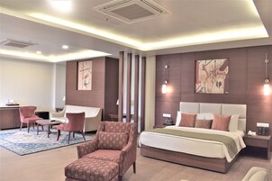 A modern hotel room with a large bed, a sitting area with a sofa and chairs, a desk, and artwork on the walls.
