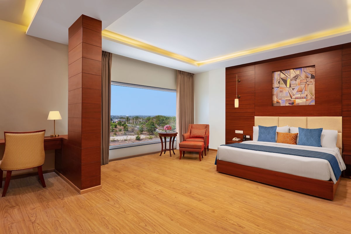 A hotel room with a large bed, a desk, a chair, a small table with a lamp, and a window with a view.