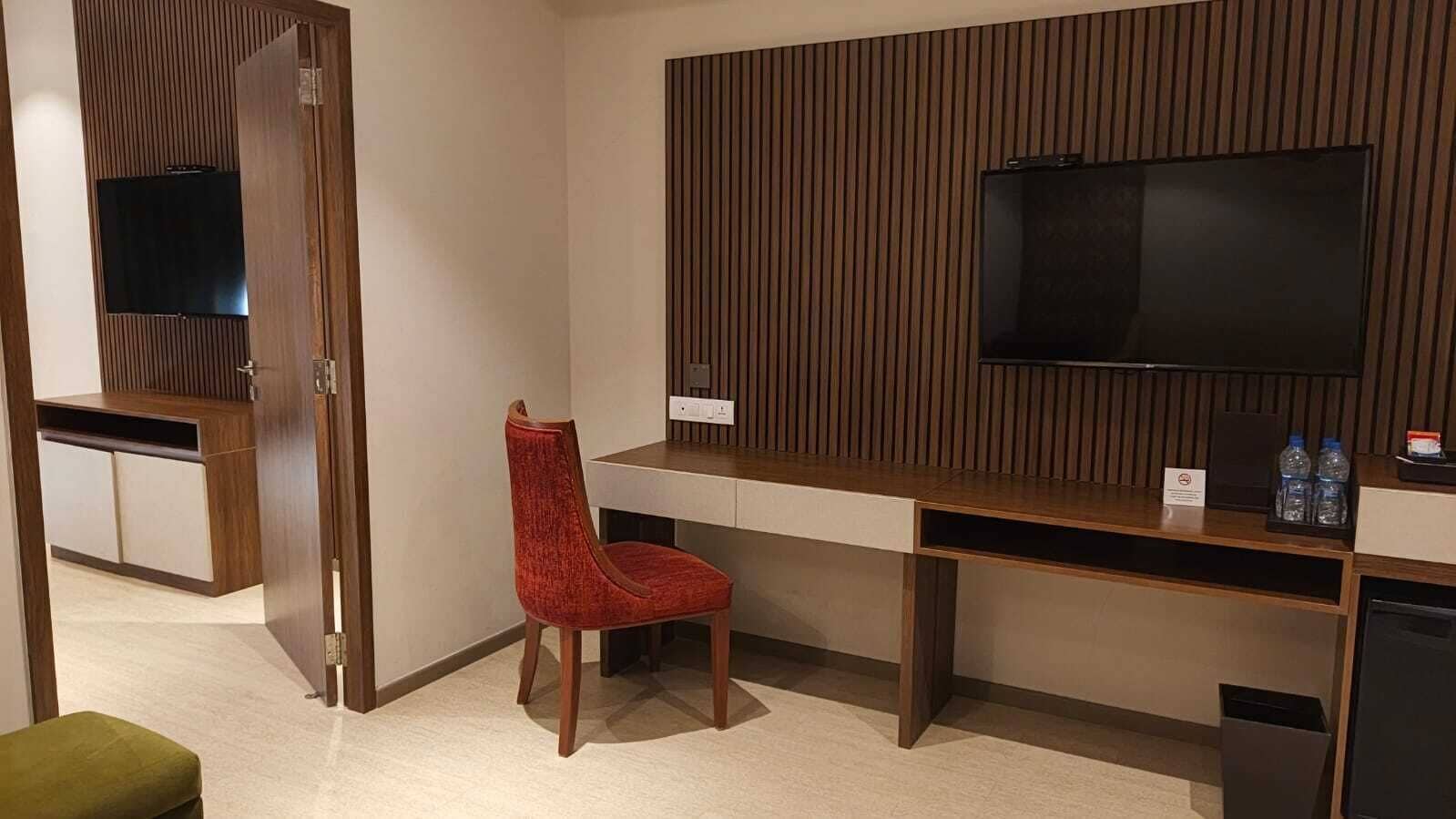 A hotel room with a wooden desk, a red chair, a television mounted on the wall, and a small table with water bottles.