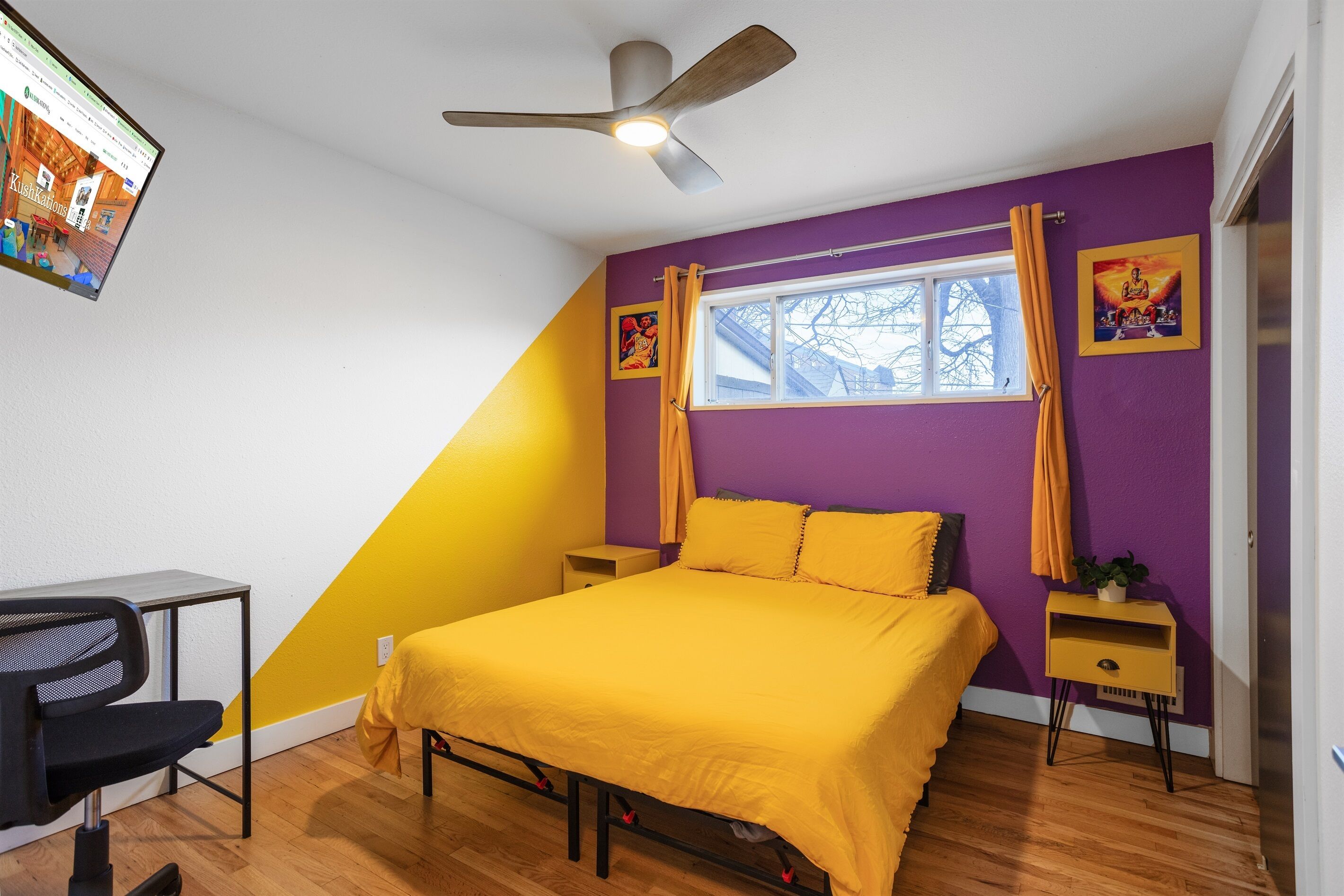 A bedroom with a bed, a desk, a chair, a ceiling fan, a window with curtains, and artwork on the walls.