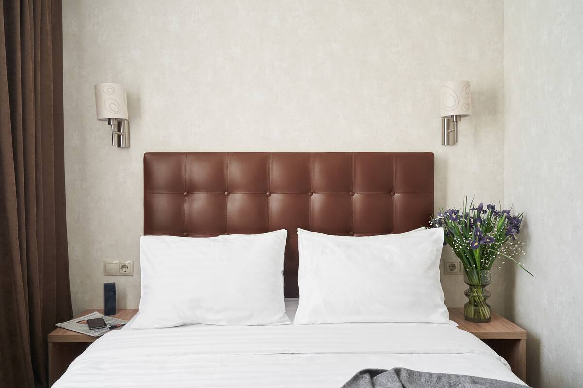 Ein Hotelzimmer mit brauner Kopfteil, weißer Bettwäsche, zwei Nachttischlampen, einem Nachttisch mit Telefon und Zeitschrift sowie einer Vase mit Blumen.