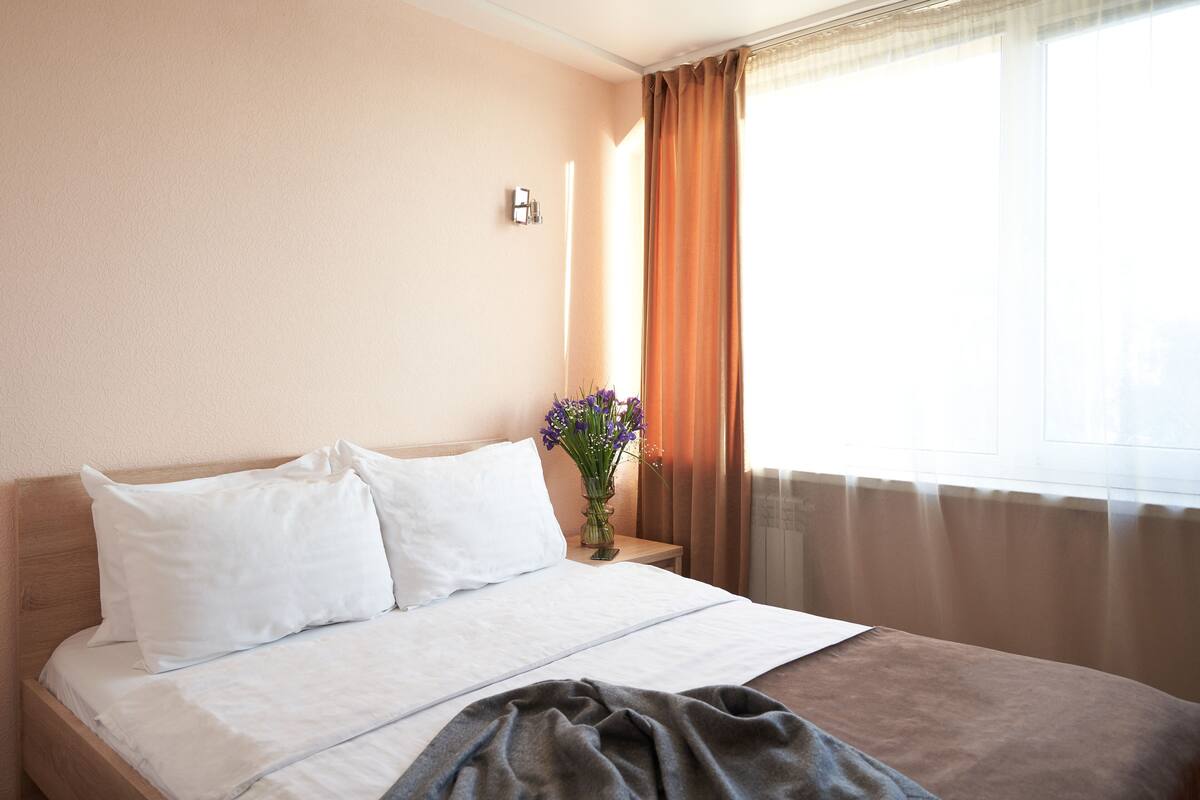 Ein Hotelzimmer mit Bett, Nachttisch, einer Blumenvase und einem Fenster mit Vorhängen.