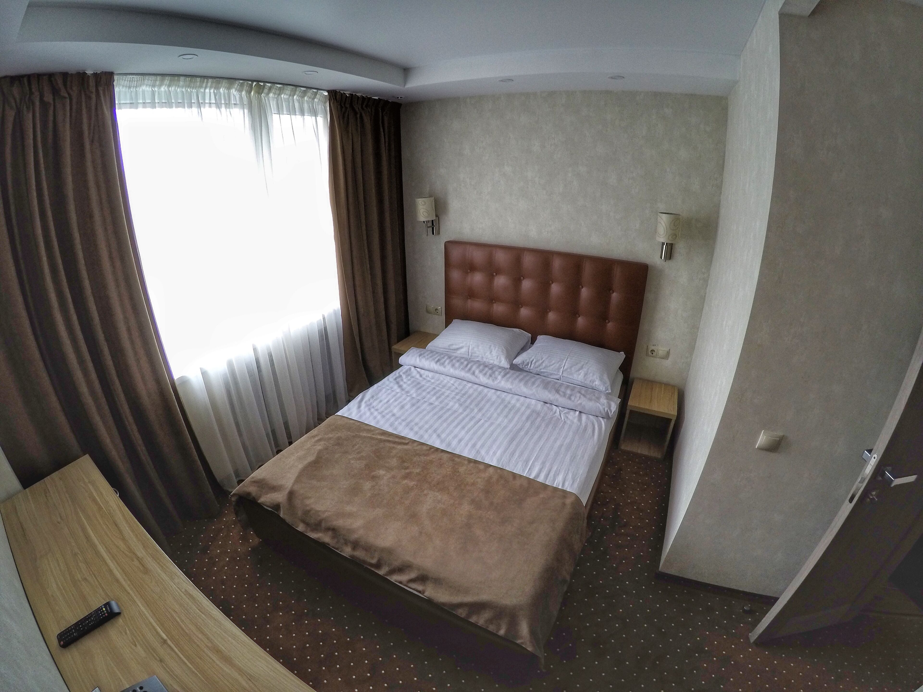 A hotel room with a bed, curtains, a nightstand, a lamp, and a remote control on a wooden table.